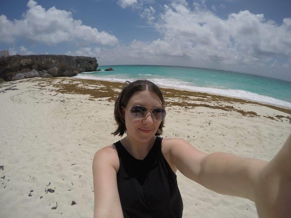 Woman taking selfie on white sand beach with seaweed and aqua water in background.