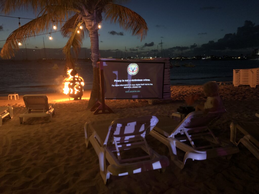 Movie screen, lounge chairs, and lit fireball on beach after sunset.