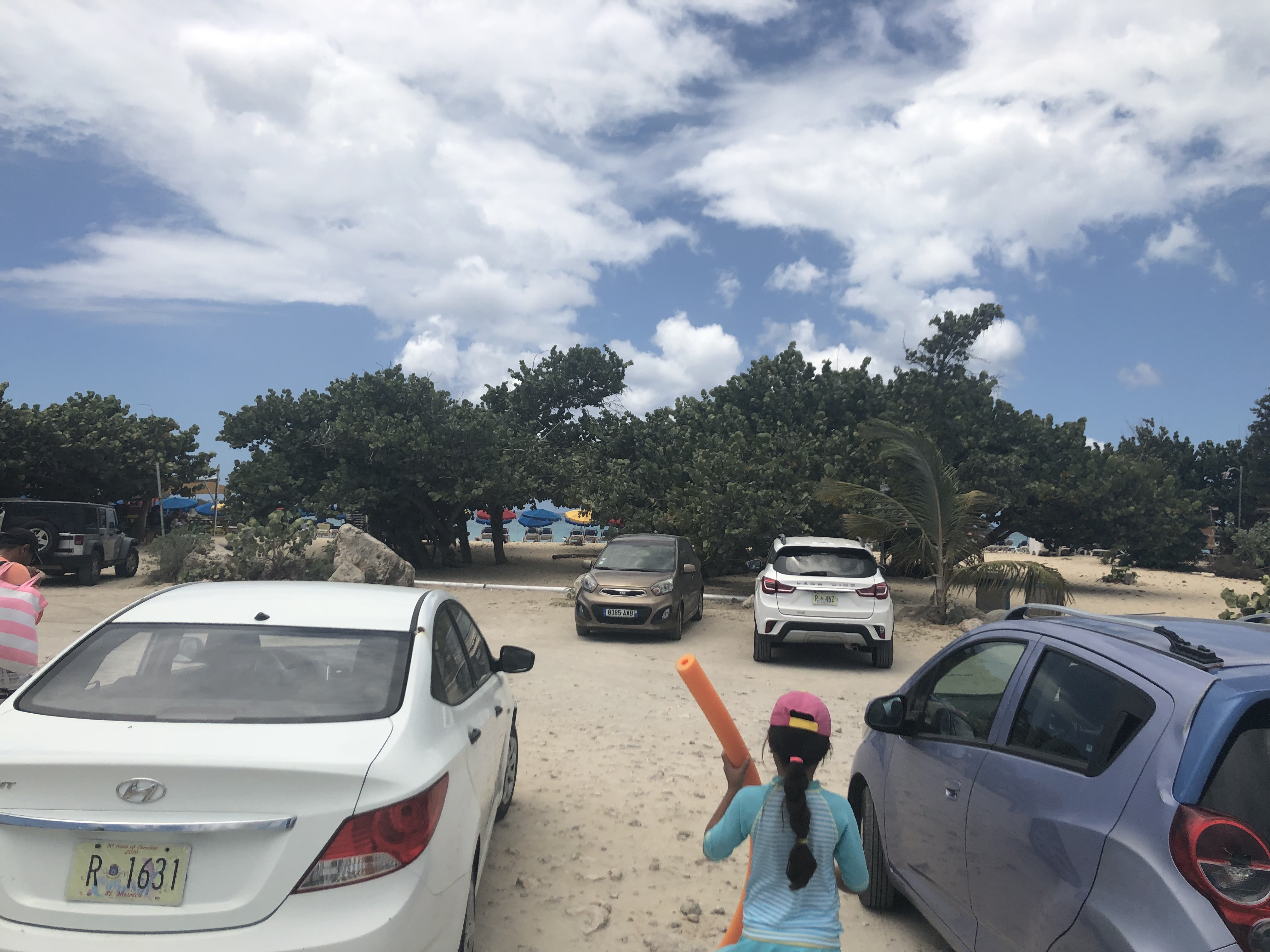 Sandy parking lot with cars and girl heading to beach.