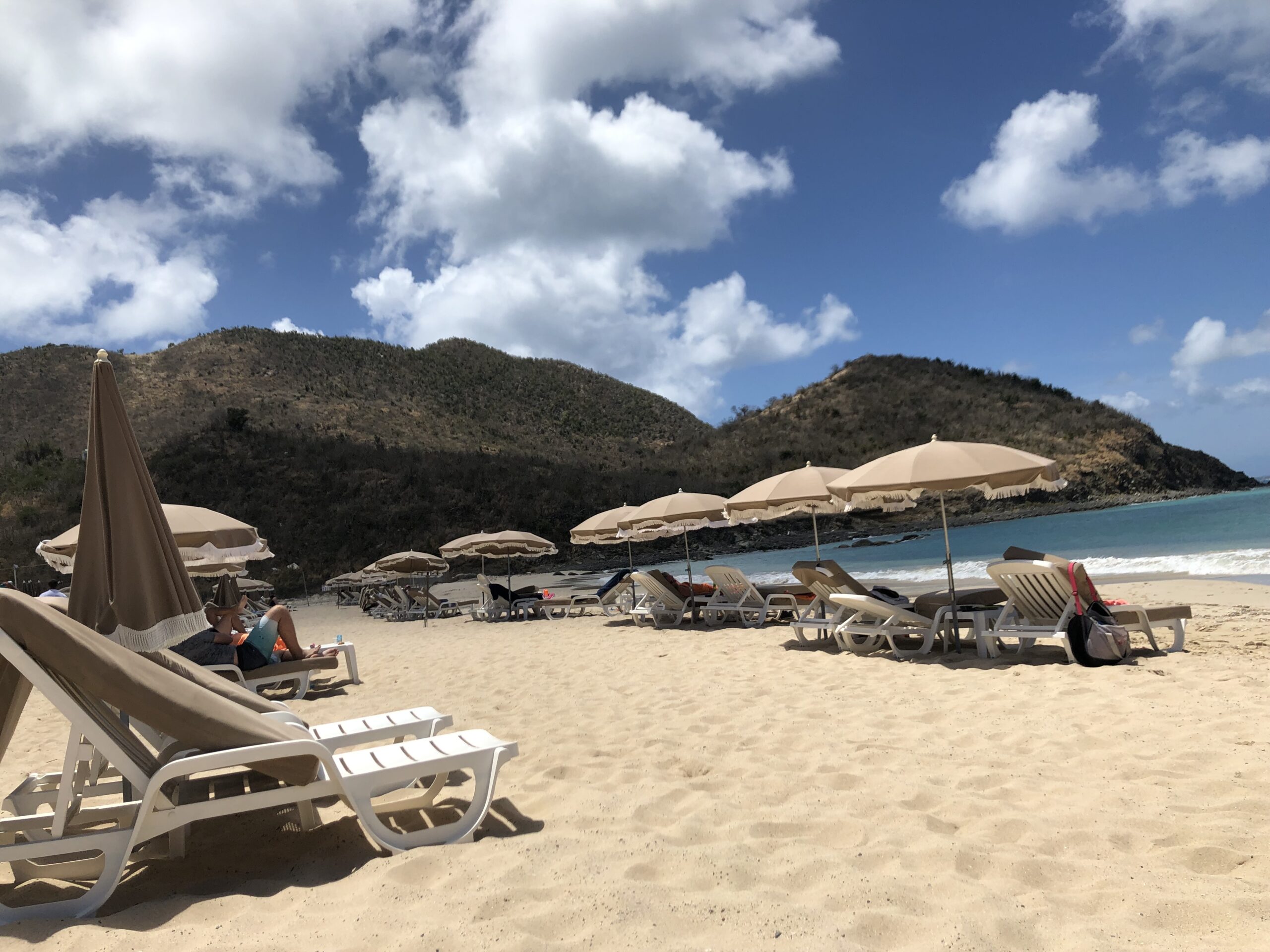 Sun beds and umbrellas set up on a beach in a cove with hills surrounding the beach.