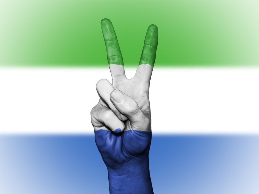 Sierra Leone flag in background with stripes of green, white and blue overlapping a hand showing the peace sign.