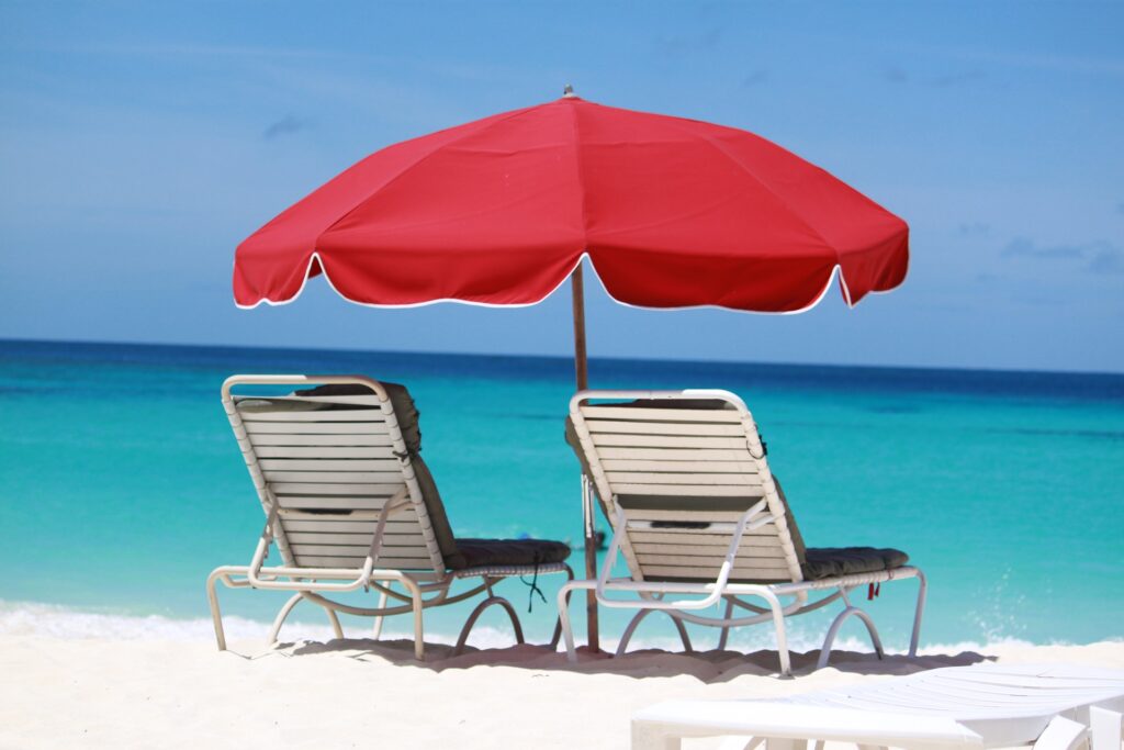 Two beach chairs beneath a red umbrella on white sand overlooking turquoise sea.