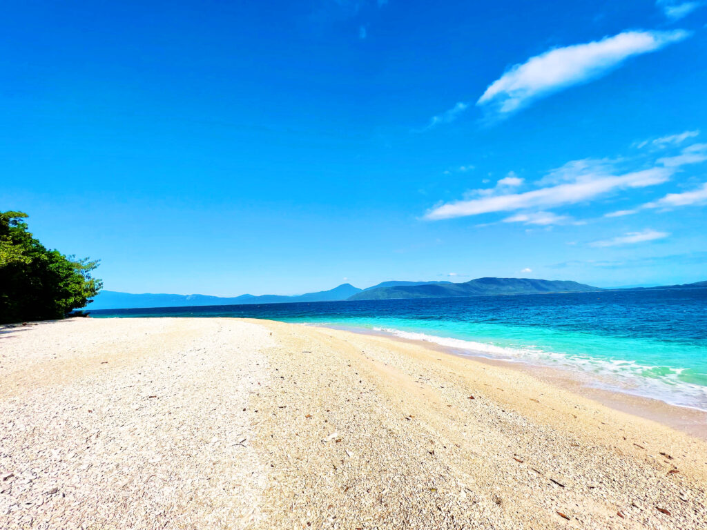 Turquoise and deep blue sea washing onto sand with green hills in distance.