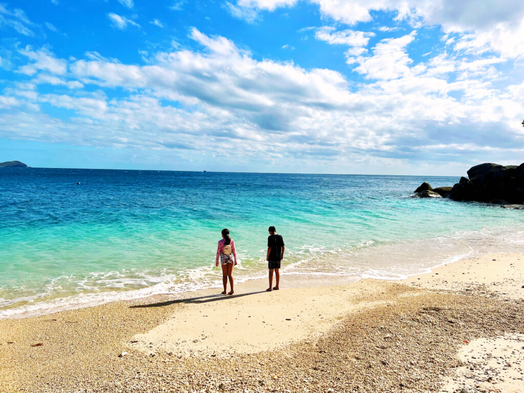 Turquoise and deep blue sea washing onto sand with two children standing at shoreline looking at water.