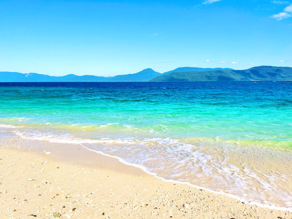 Turquoise and deep blue sea washing onto sand with green hills in distance.