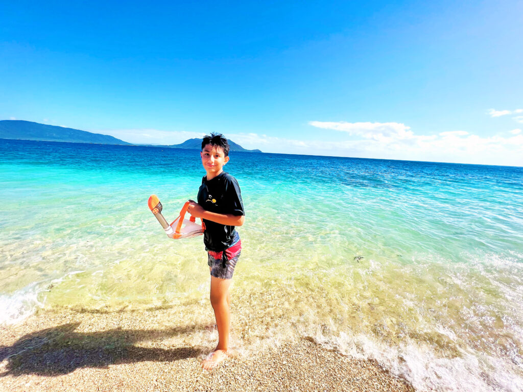 Boy holding snorkel standing on shoreline with turquoise and deep blue sea behind him.