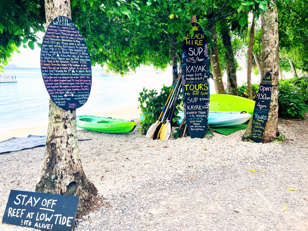 Bright colored writing on surfboard shaped chalkboards leaning against tree trunks next to a beach.