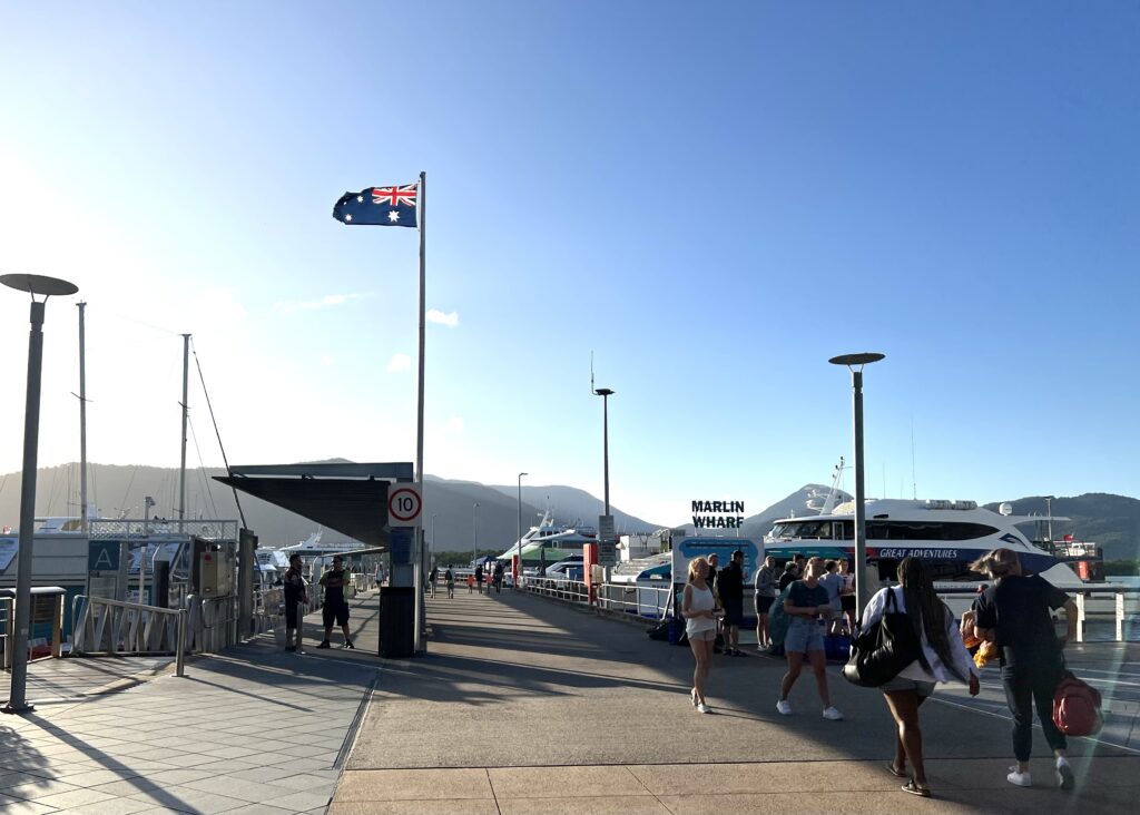 Australian flag blowing in the wind on a pole attached to the Marlin Wharf with boats docked and people walking.