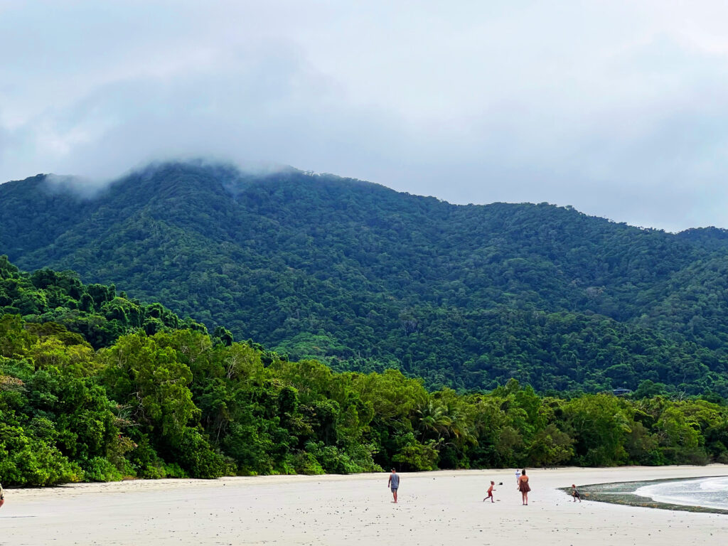 Looking across a wide beach towards hills covered in green jungle with whisps of clouds obscuring the hilltops.