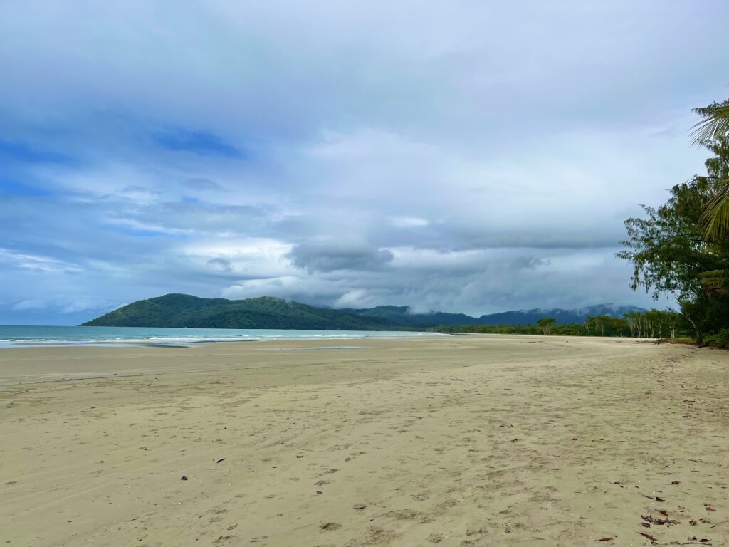 Wide beach with ocean on left and hilly rainforest on right.