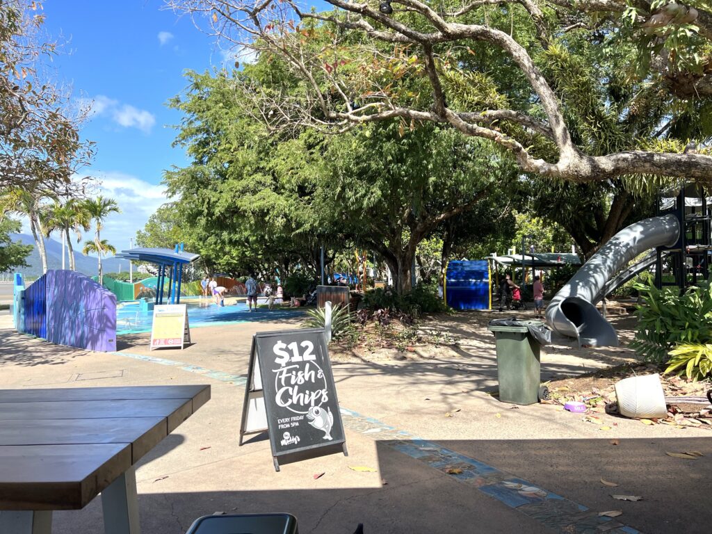 An outdoor splash park and playground beneath large trees.