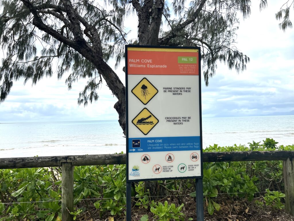 Sign warning of crocodiles and dangerous sealife next to tree overlooking beach.