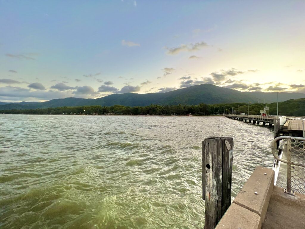 Sunset from a jetty looking back at beach and green hills beyond.