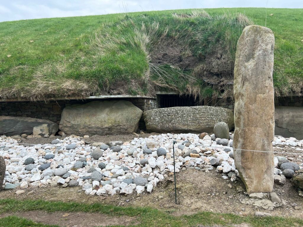 Entrance with iron bars blocking it beneath a green mound surrounded by boulders at the bottom.