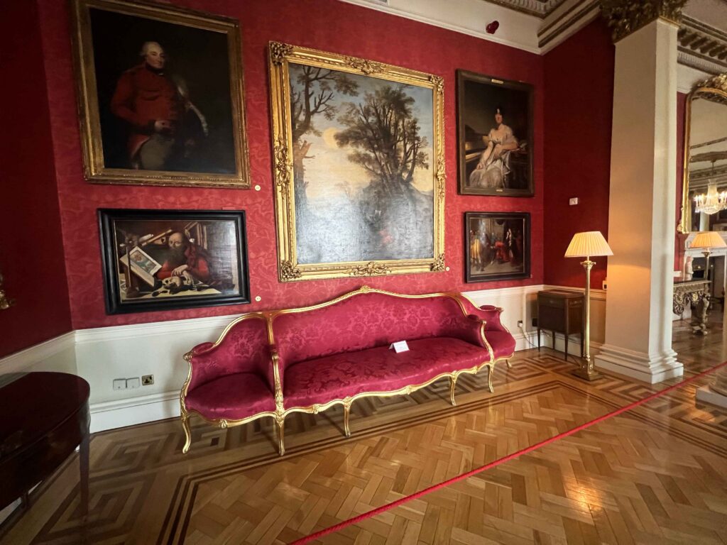 Ornate red velvet couch set before a wall displaying gold framed old paintings.