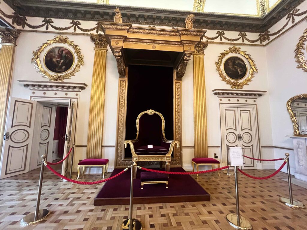 An ornate velvet and gold throne set on a raised platform in a room with white walls and ornate golden carvings.