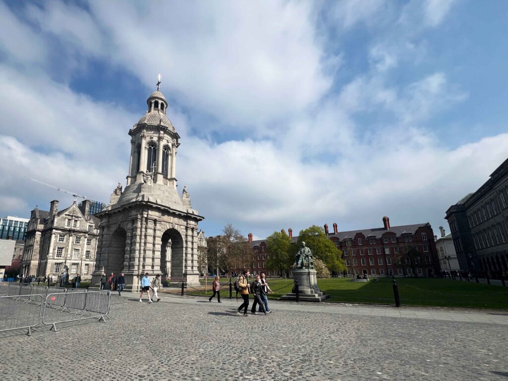 Stone and brick campus buildings surrounding a green space with students walking past.