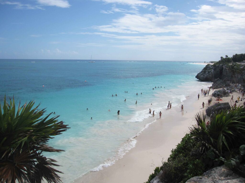 Narrow white sand beach next to rocky cliffs with swimmers in the aqua ocean water.